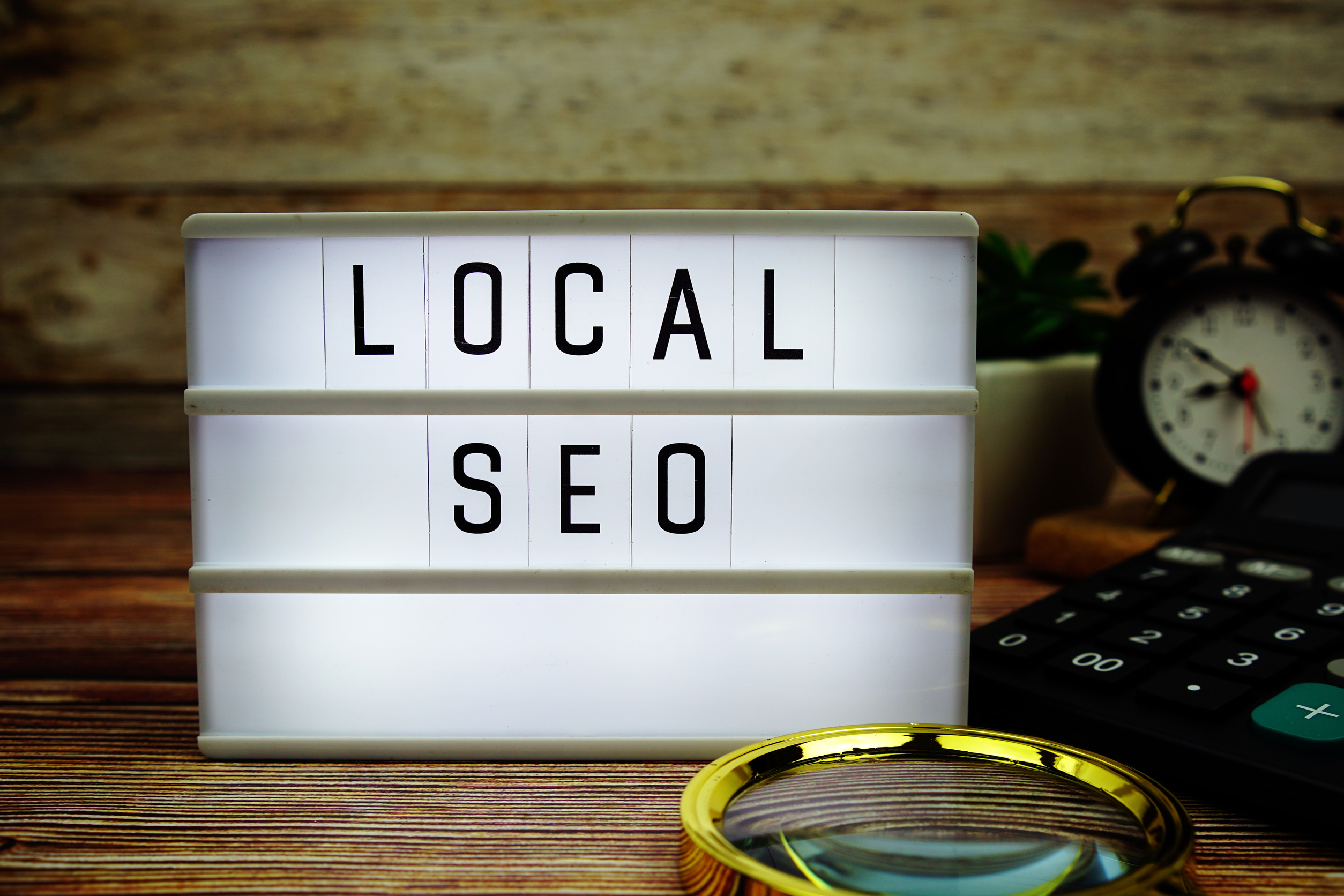 A lightbox with the words "LOCAL SEO" sits on a wooden table, next to a magnifying glass, calculator, potted plant, and an alarm clock—perfect for showcasing local SEO or Google Business Profile optimization strategies.