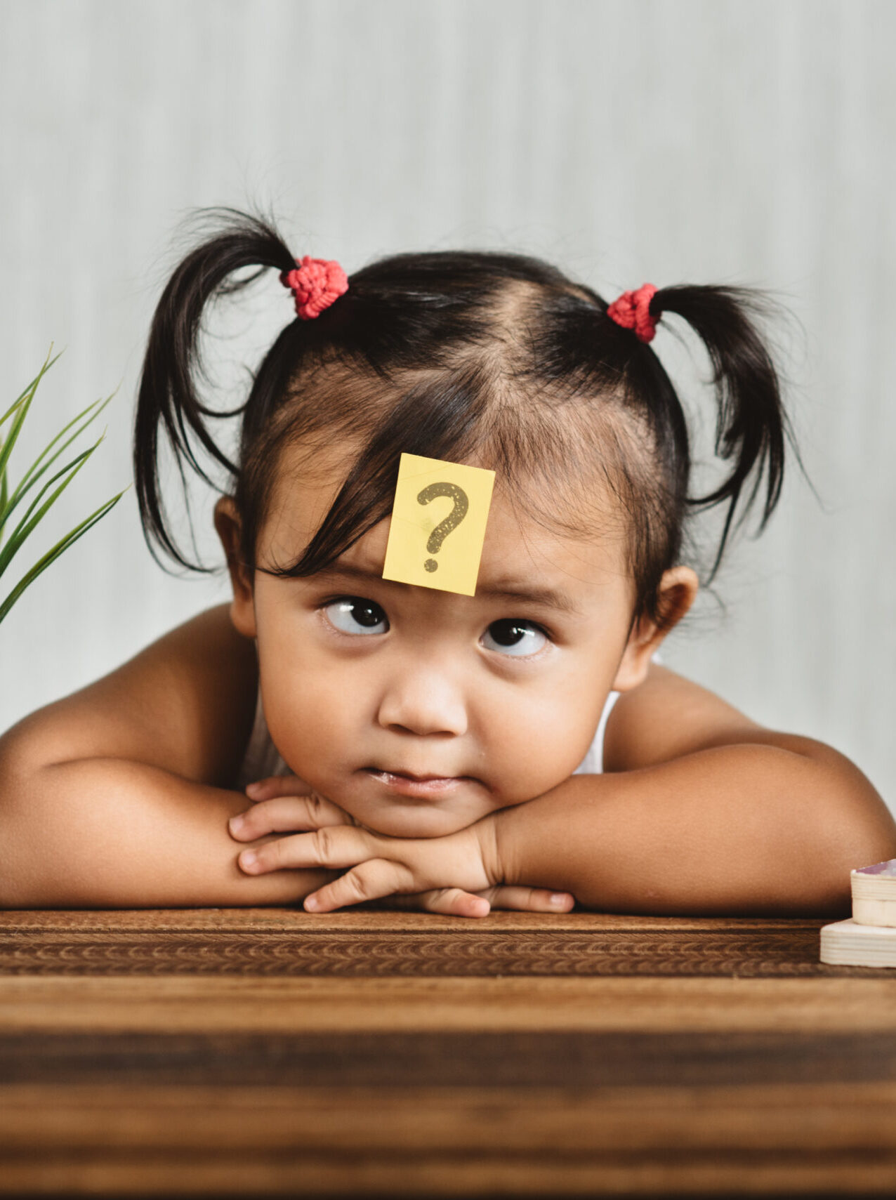 A young child with pigtails rests her chin on her arms at a table, with a yellow sticky note featuring a question mark on her forehead. A potted plant, stack of books, and alarm clock are on the table.