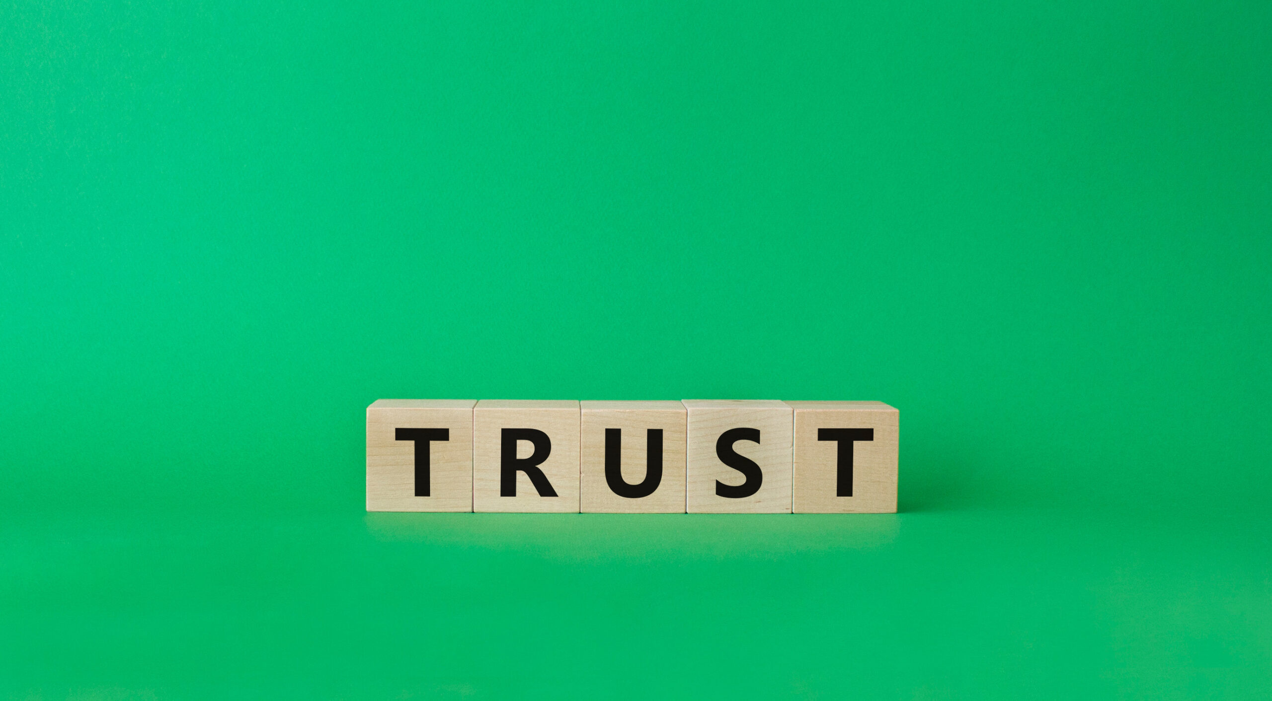 Wooden blocks spelling the word "TRUST" are arranged in a row against a plain green background.