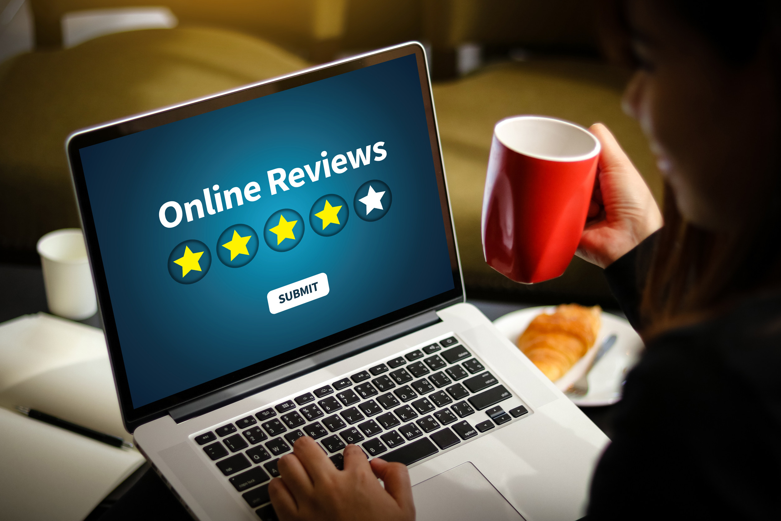 A person holding a red mug uses a laptop displaying "Online Reviews" with a four out of five star rating and a "Submit" button, highlighting the impact of online reviews on local SEO. A croissant rests on the table nearby.