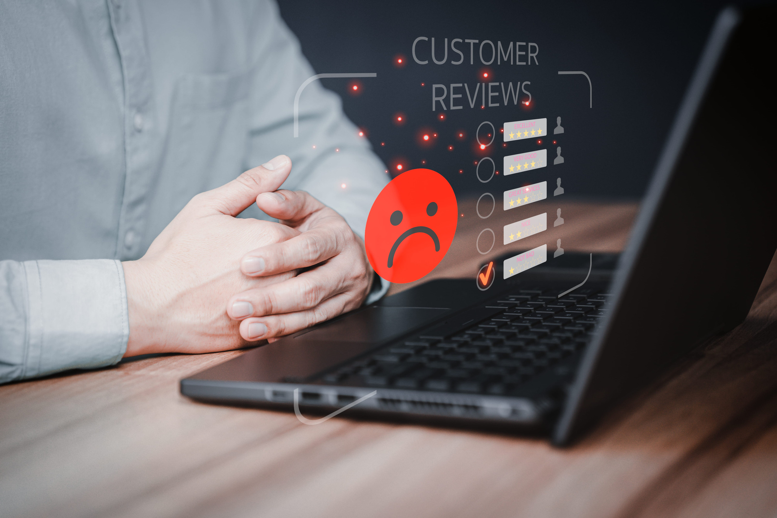 A person sits at a desk with hands clasped near a laptop displaying a virtual screen showing "Customer Reviews" and a large red sad face, indicating negative feedback.