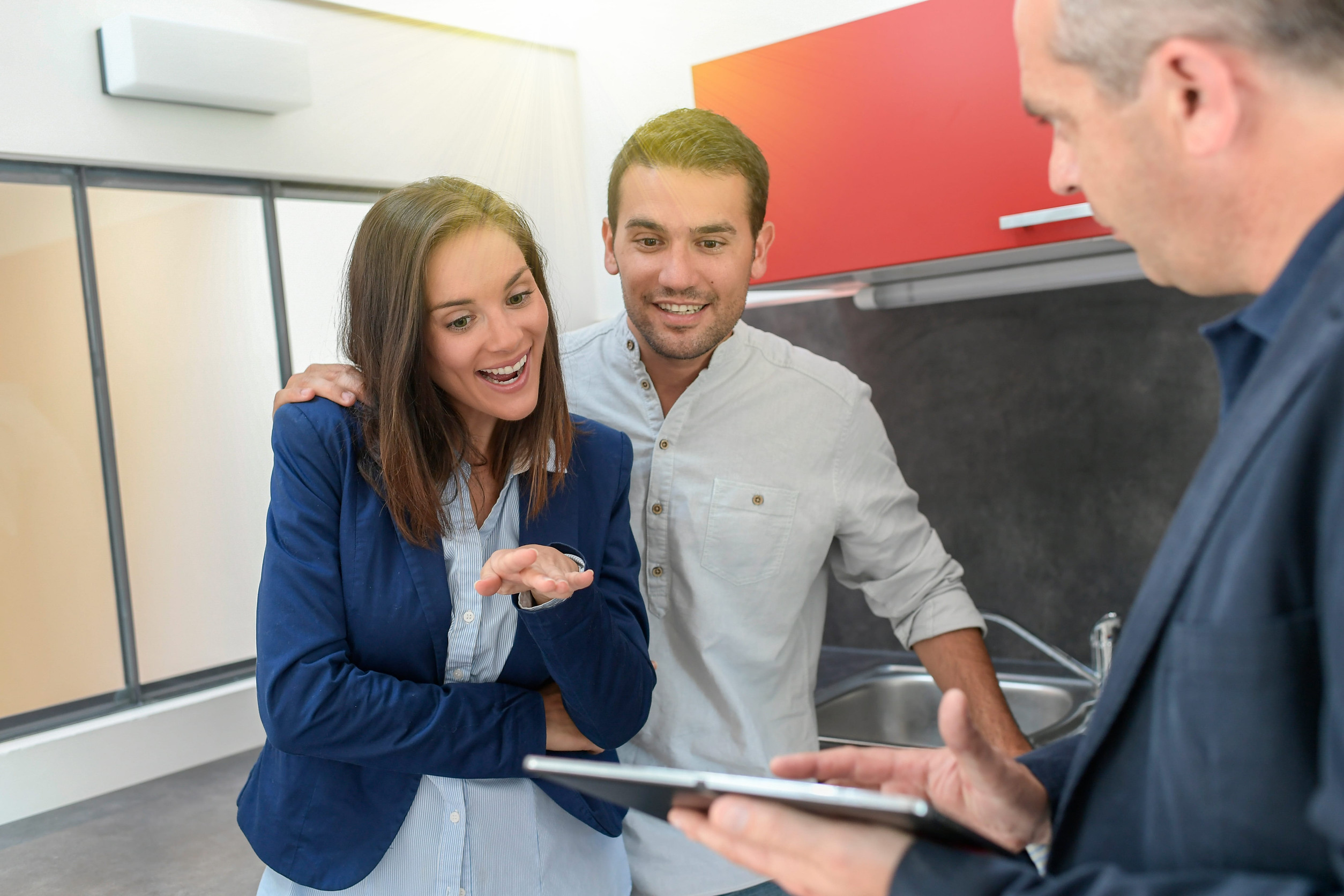 A smiling couple stands in a modern kitchen, looking at a tablet held by a man in a suit. They appear engaged and happy, possibly discussing customer reviews about their real estate experience, all without sounding pushy.